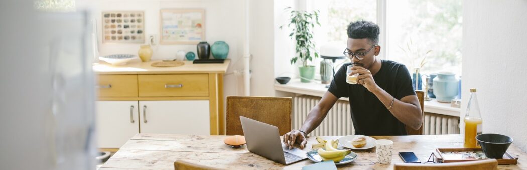 Young man drinking juice while using laptop at table in house