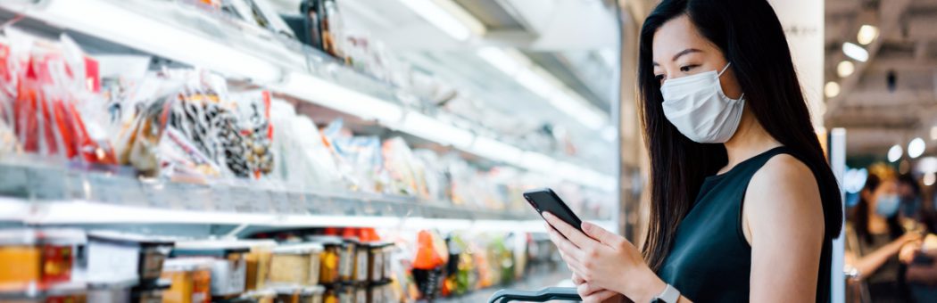 Young woman with protective face mask holding shopping basket and using smartphone while grocery shopping in a supermarket