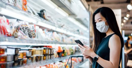 Young woman with protective face mask holding shopping basket and using smartphone while grocery shopping in a supermarket