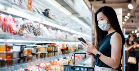 Young Asian woman with protective face mask holding shopping basket and using smartphone while grocery shopping in a supermarket