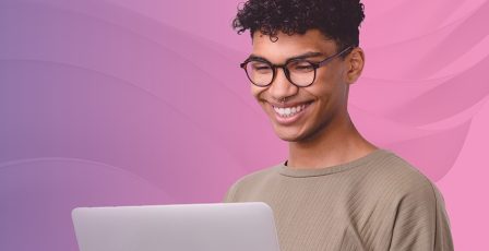 Person smiling while using a laptop, stood in front of a pink and purple gradient