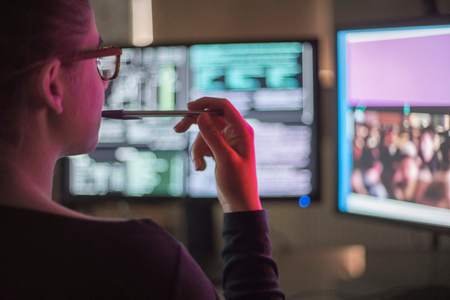 Stock image of a young woman contemplating two computer monitors, she is illuminated by the red light from a peripheral screen image.