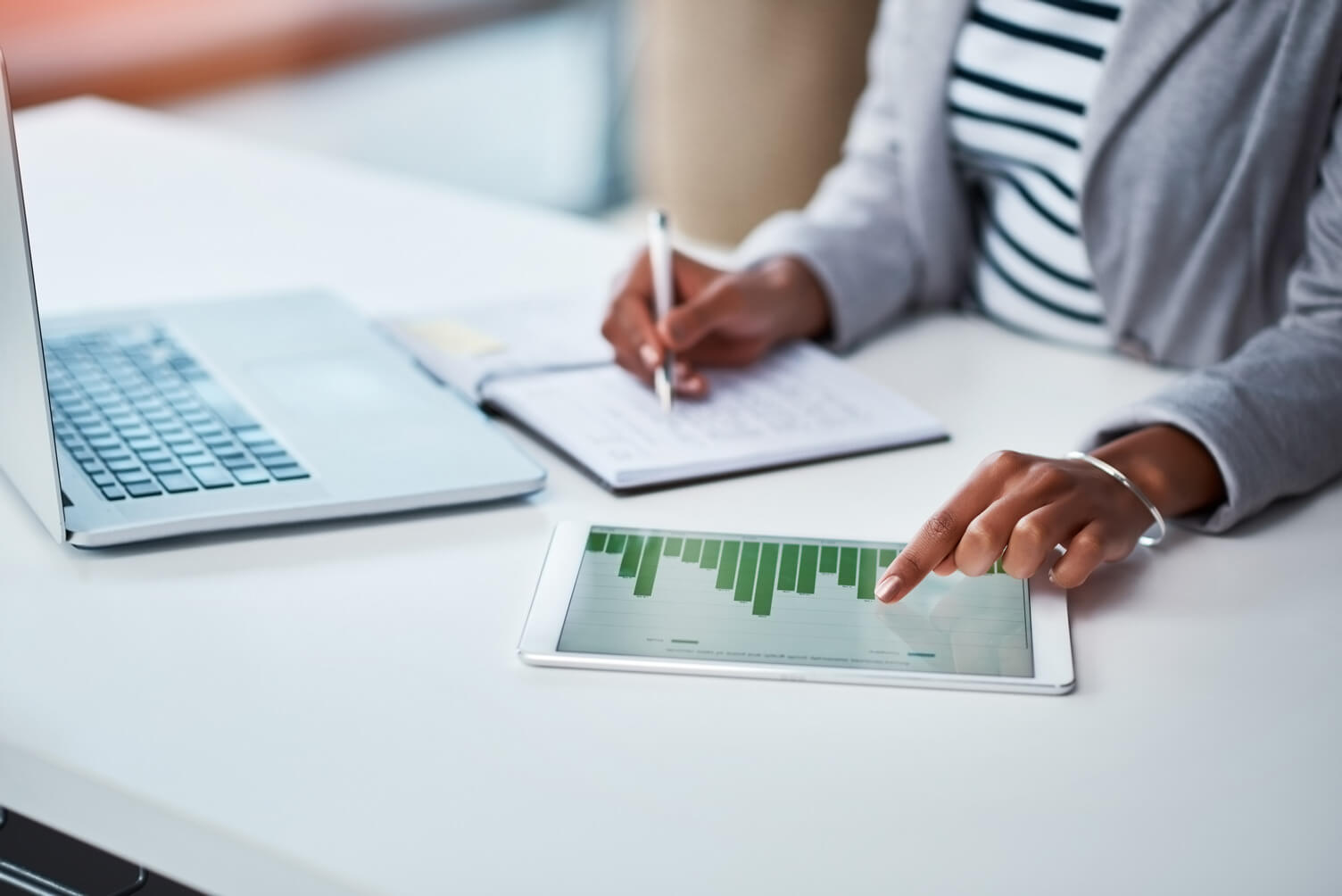 Woman's hands pointing at a table chart on a tablet and the other writing notes in a notepad