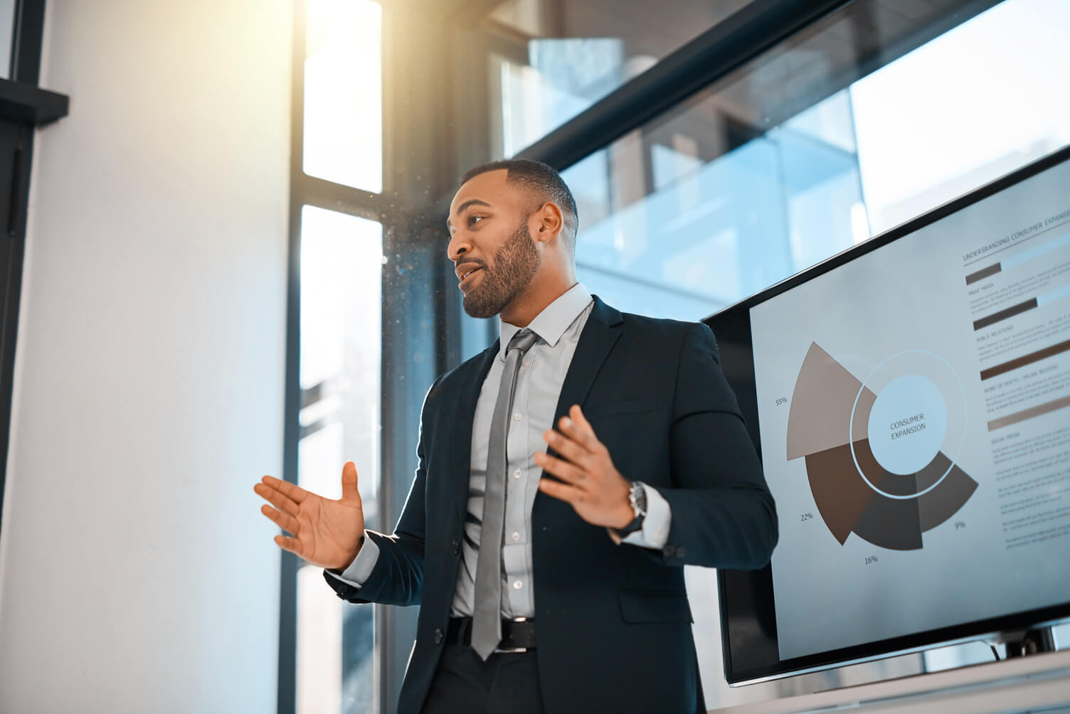 A man talking in front of a large tv screen with data on